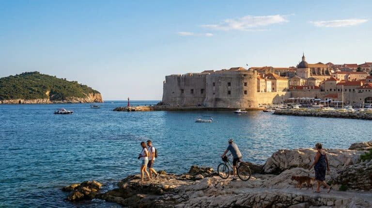 The fortified walls of Dubrovnik's Old Town seen from the rocky Adriatic coast with people walking and cycling.