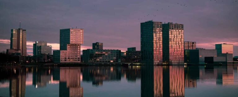 The modern buildings of the Almere city center skyline reflecting in calm water during a colorful dusk.