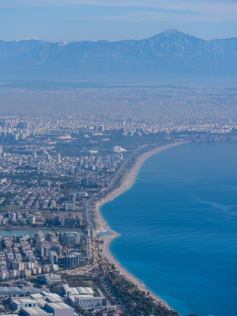 An expansive aerial view over the city of Antalya, with Konyaaltı Beach meeting the blue sea and the Taurus Mountains in the background.