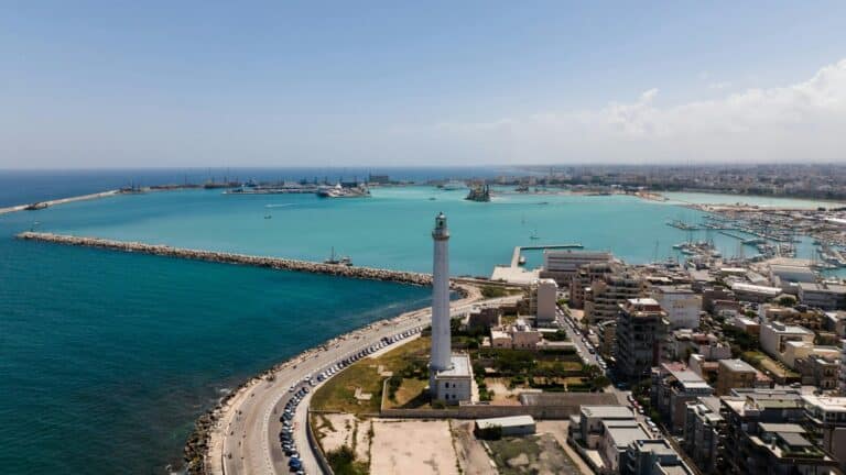 An aerial view of Bari, Italy, featuring the Faro di San Cataldo lighthouse overlooking the turquoise waters of the port.