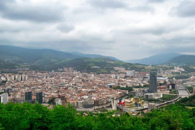 A panoramic view of the Bilbao cityscape, with the Guggenheim Museum and the Nervión River surrounded by green hills.