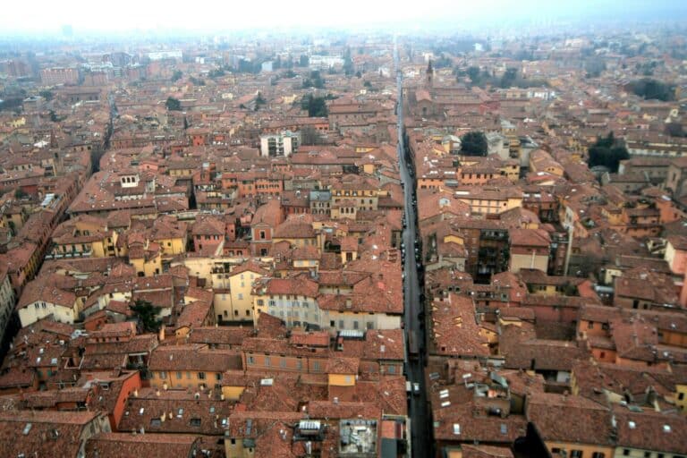 A high-angle view over the dense, terracotta-tiled roofs of Bologna's historic center on a hazy, overcast day.