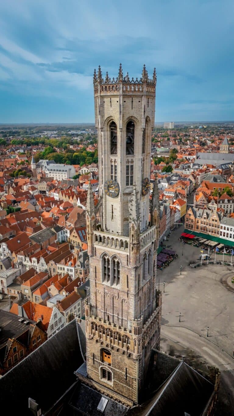 An aerial view of the medieval Belfry of Bruges rising above the historic city center's red-roofed buildings.