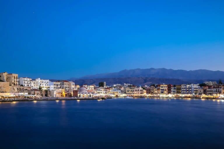 The Old Venetian Harbour in Chania, Greece, is illuminated by city lights against a deep blue twilight sky.