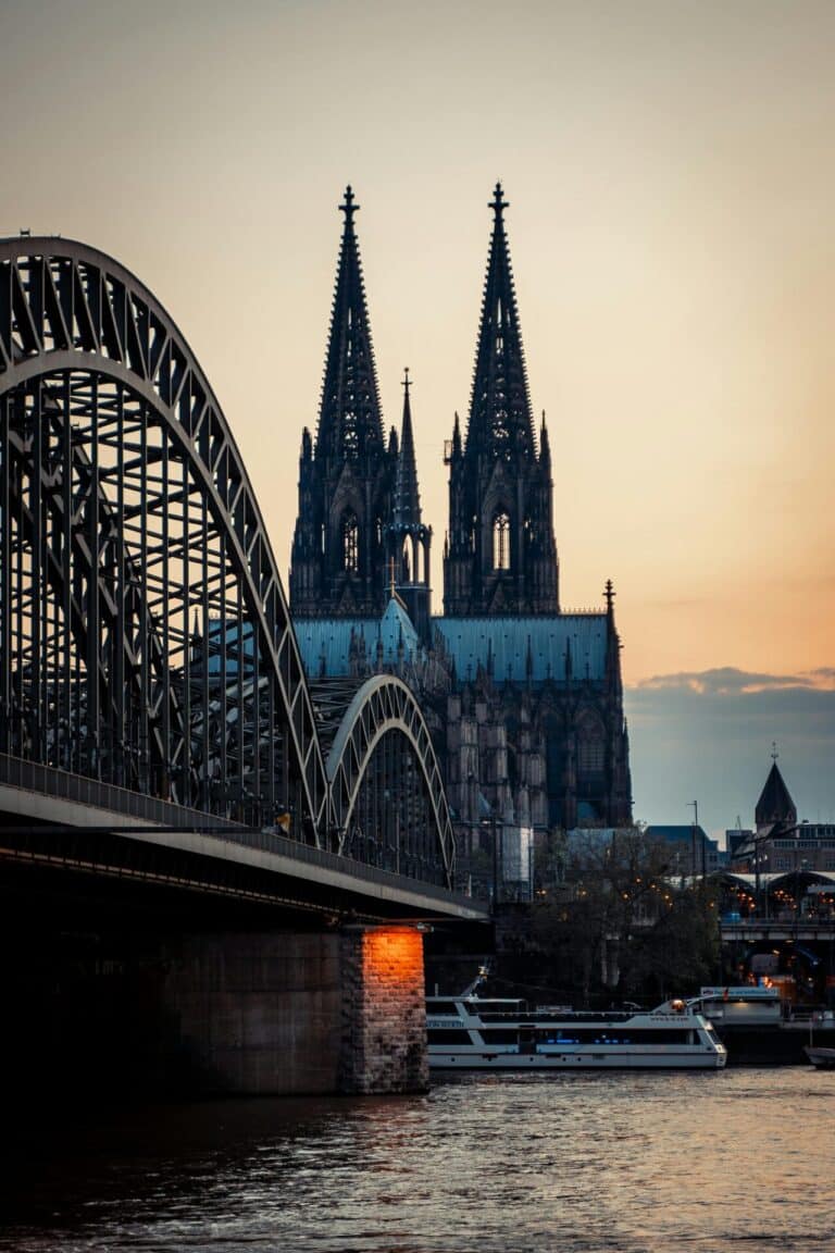 The Hohenzollern Bridge over the Rhine River with the twin spires of the Cologne Cathedral at dusk.