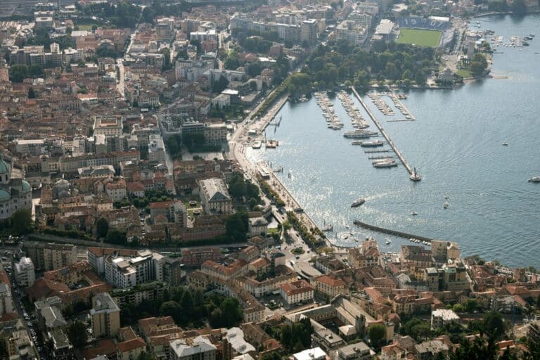 An aerial daytime view of Como, Italy, overlooking the city's historic buildings, waterfront, and the sparkling blue Lake Como.