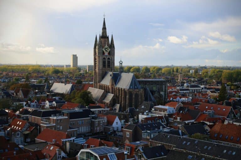 An aerial view over the rooftops of Delft, focusing on the historic Oude Kerk rising above the city.