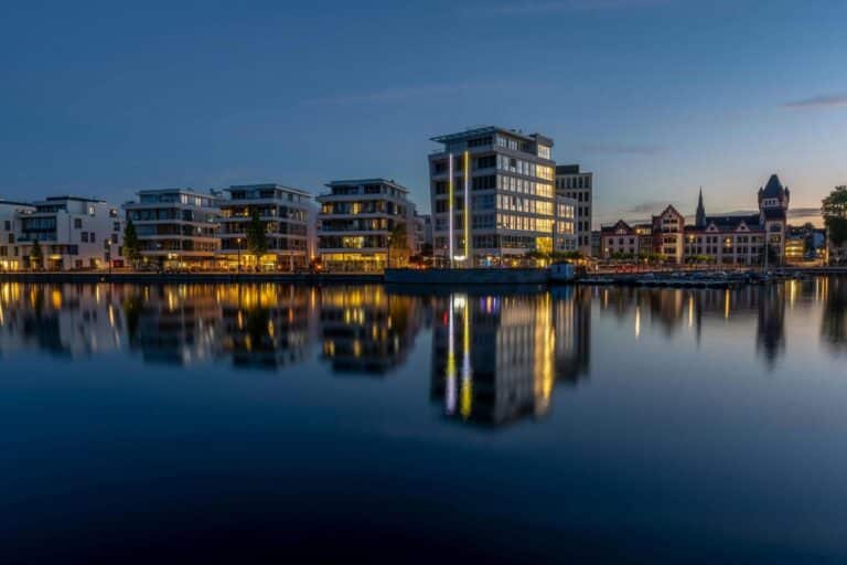 Modern buildings and historic architecture are reflected in the calm waters of Phoenix See in Dortmund at dusk.