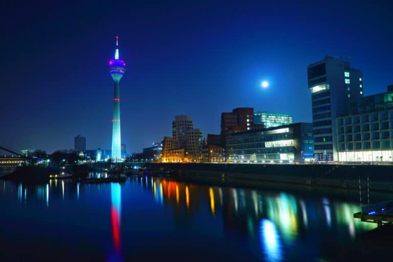 The illuminated Rheinturm tower and Medienhafen buildings in Düsseldorf at night, with reflections in the Rhine river.