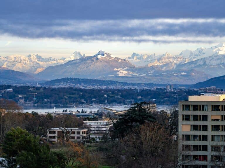 An elevated view over the city of Geneva, with Lake Geneva and the snow-covered Alps in the background.