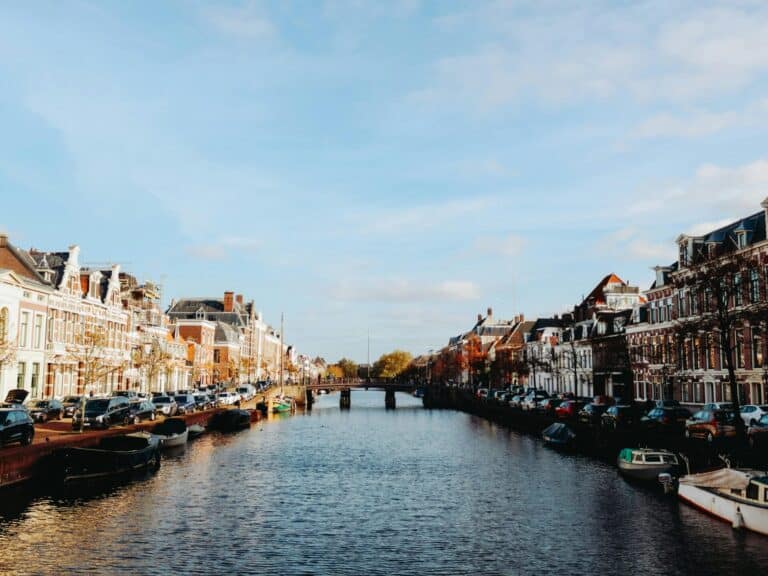 A scenic view down the Nieuwe Gracht canal in Haarlem, lined with historic Dutch houses, parked cars, and boats.