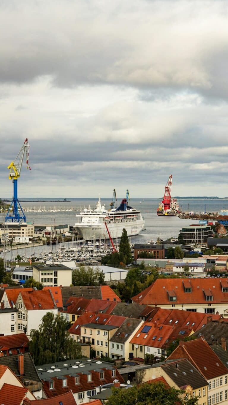 An elevated view over the red-tiled rooftops of Kiel, Germany, looking towards the bustling harbor with a cruise ship.