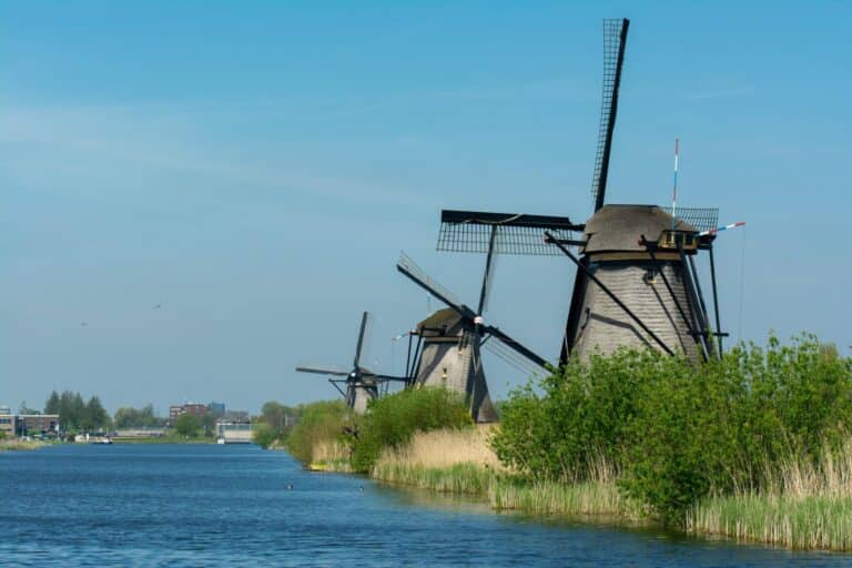 A row of historic Dutch windmills with thatched roofs lining a tranquil canal in Kinderdijk under a clear blue sky.