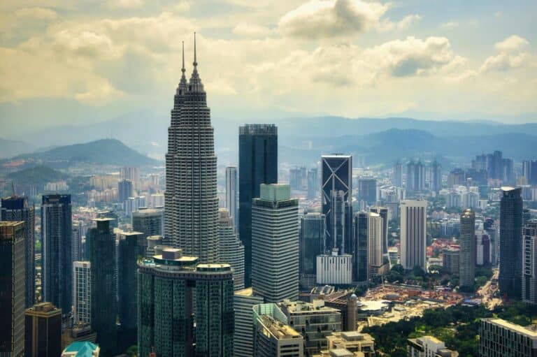 An aerial view of the Kuala Lumpur skyline, featuring the Petronas Twin Towers among many other skyscrapers with mountains behind.