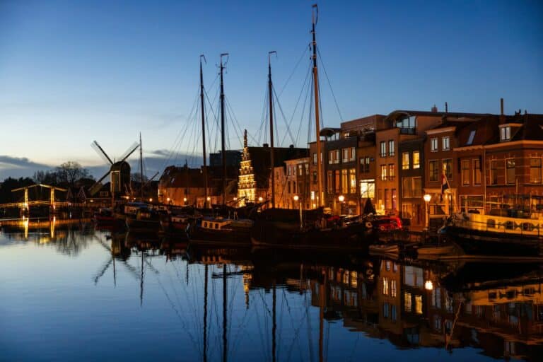 Historic sailing ships moored in the Oude Haven of Leiden at dusk, with an illuminated windmill in the background.