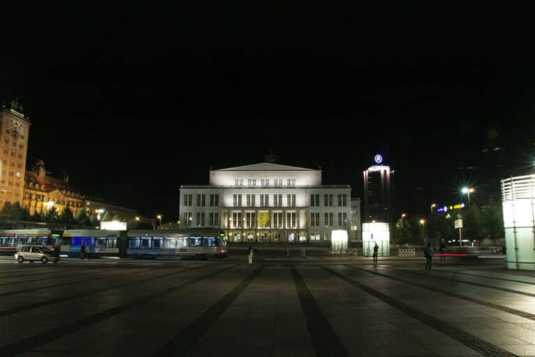 The brightly illuminated Leipzig Opera House on Augustusplatz at night, with trams and pedestrians in the large public square.