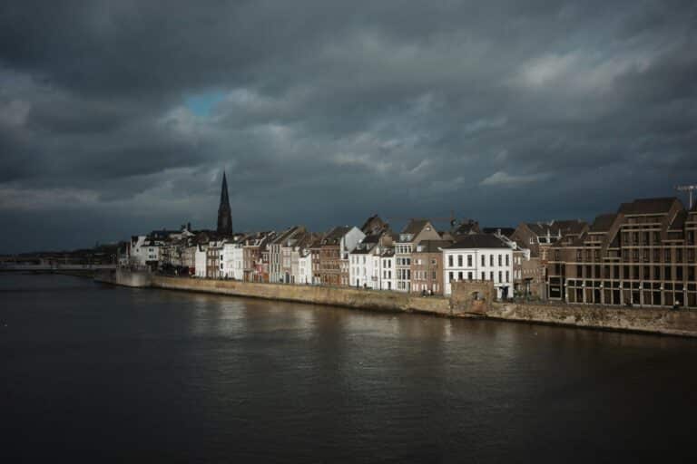 The historic waterfront of Maastricht's Wyck district seen from across the Meuse river on a dramatic, overcast day.