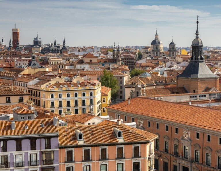 An aerial view over the historic terracotta rooftops and church spires of central Madrid on a partly cloudy day.