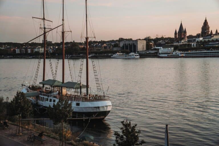 A three-masted ship docked on the Rhine River in Mainz, Germany, with the city skyline and cathedral at dusk.