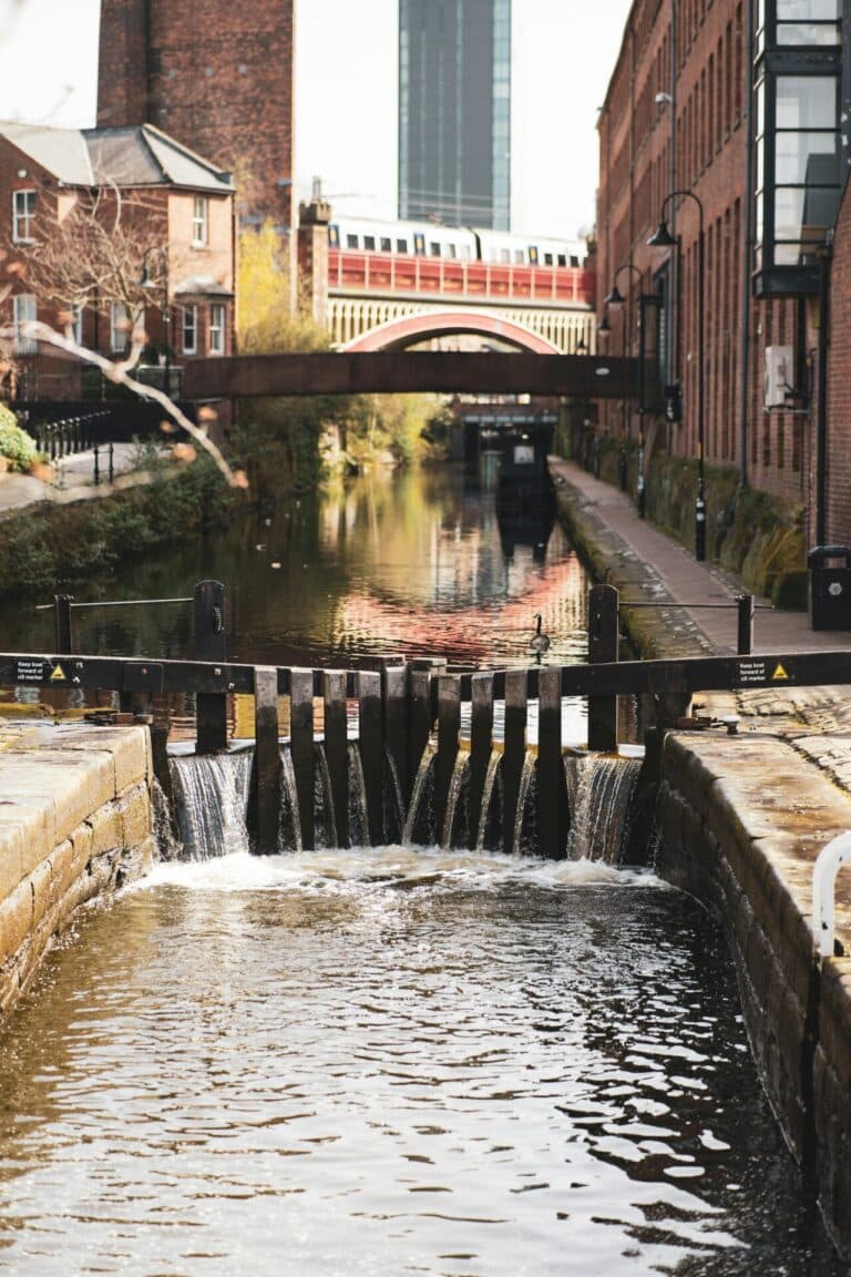 Water flows over a wooden lock gate on a canal in Manchester, surrounded by old red brick buildings.