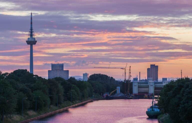 The Fernmeldeturm tower in Mannheim stands against a colorful purple and pink sunset sky above a canal.