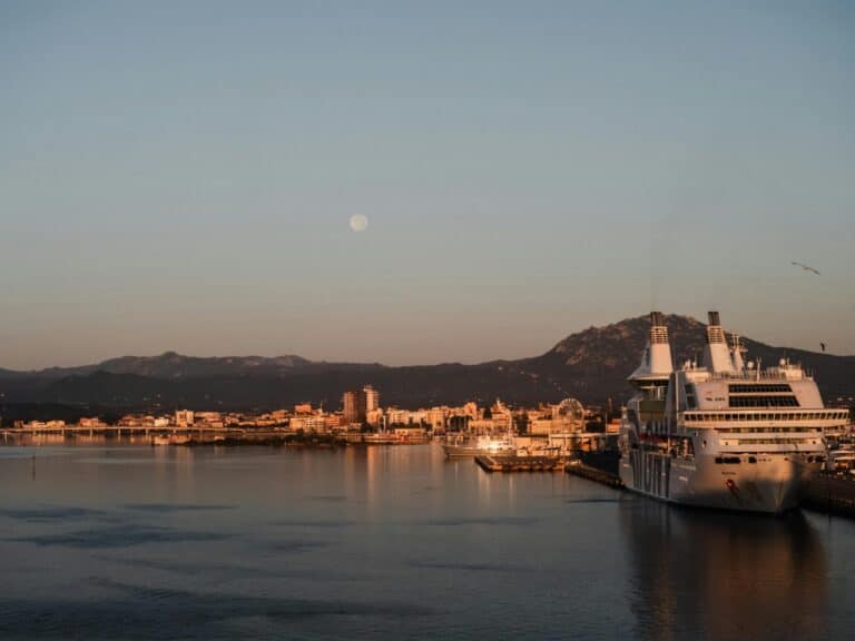The port of Olbia at dusk with a large white ferry docked and the moon visible over the city.