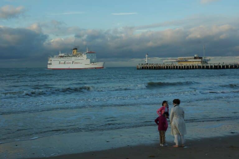 A woman and two children stand on the beach in Ostend watching a large ferry sail past the pier.