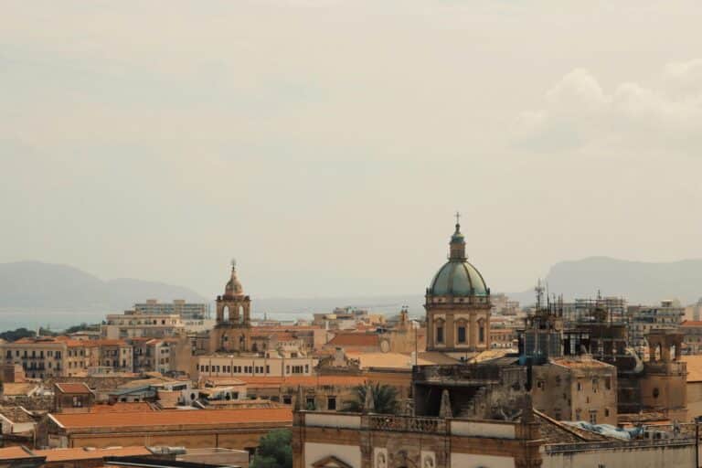 A hazy view over the historic rooftops and church domes of Palermo, Italy, with mountains in the distance.