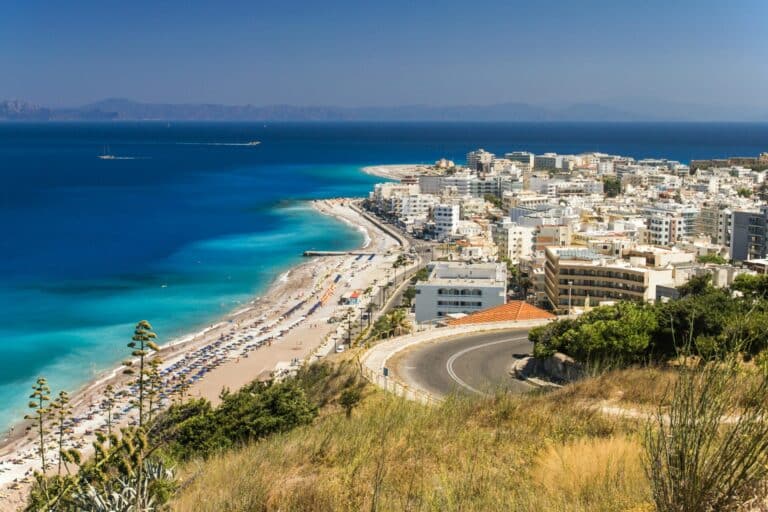 An elevated daytime view of the city of Rhodes, showing the long beach, turquoise water, and city buildings.