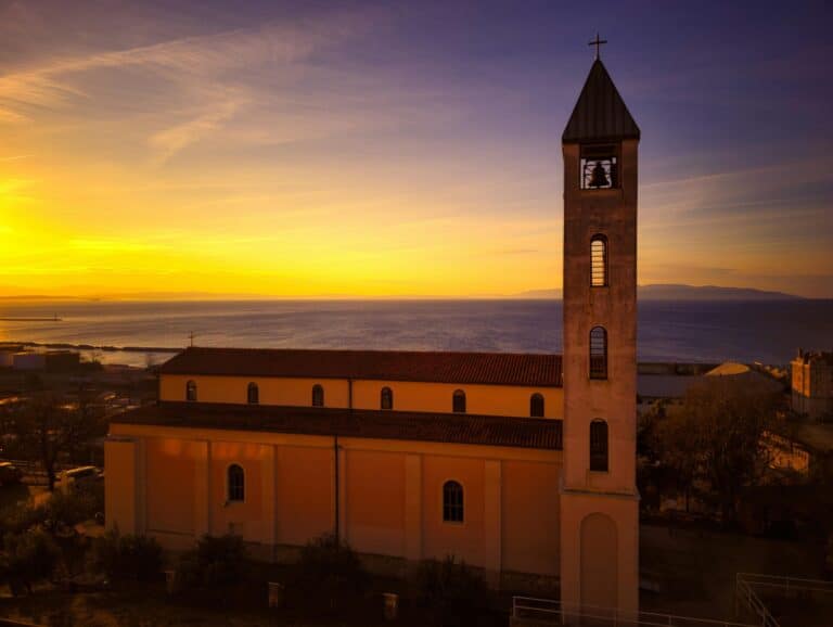 The Church of St. Joseph in Rijeka, Croatia, with its prominent bell tower overlooking the sea during a golden sunset.