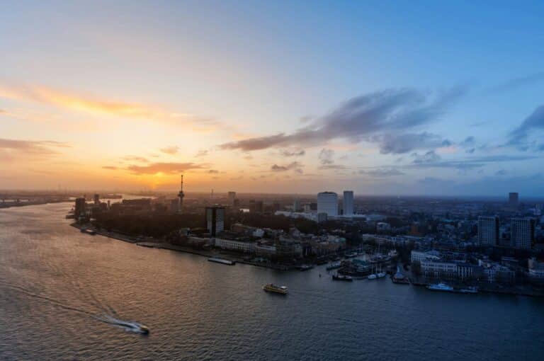 An expansive aerial view of the Rotterdam skyline, the Euromast tower, and the Nieuwe Maas river at sunset.