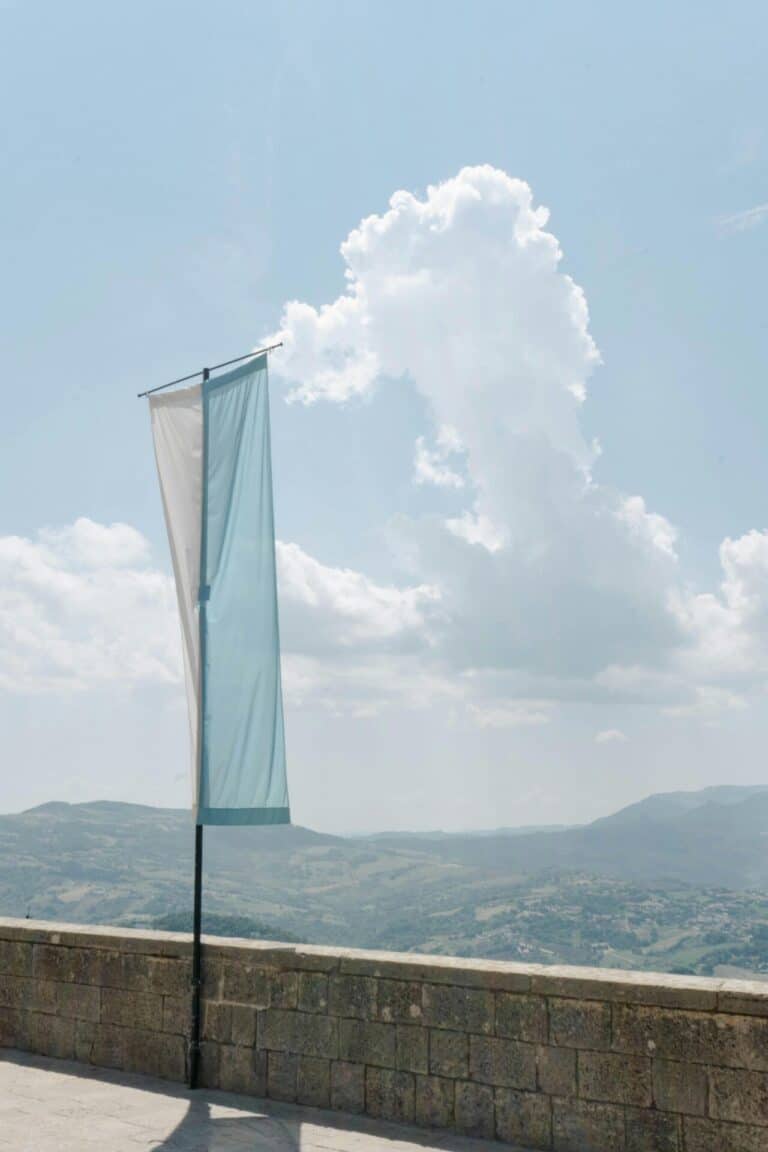 The flag of San Marino flies on a pole from a stone wall overlooking the country's rolling green hills.