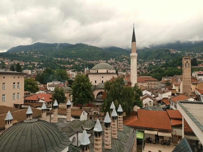 An elevated view over the historic rooftops of Sarajevo, showing the Gazi Husrev-beg Mosque and clock tower.