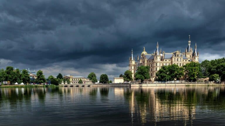 Schwerin Palace sits on the shore of a lake, its reflection visible in the water under dramatic storm clouds.
