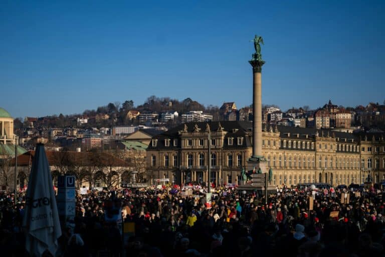 A large crowd protests in Stuttgart's Schlossplatz with the New Palace and Jubilee Column under a clear sky.
