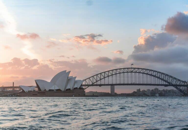 The Sydney Opera House and Harbour Bridge are seen from the water with a colorful sunset sky.