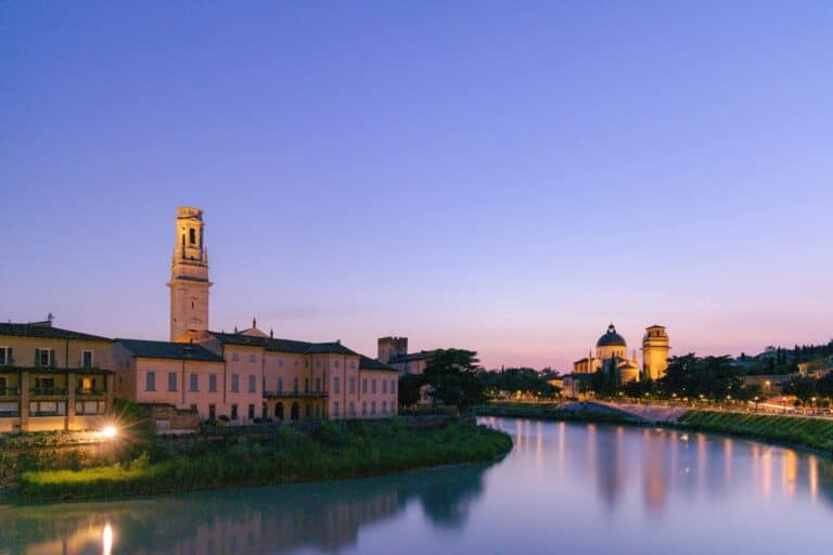 The Adige River in Verona at dusk, showing the illuminated Verona Cathedral bell tower and historic buildings.