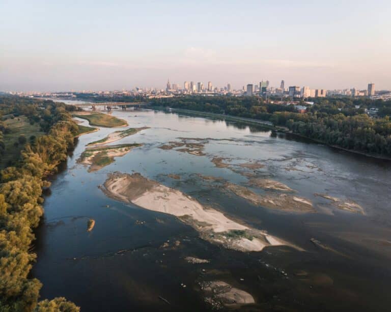 Aerial view of the Vistula River in Warsaw showing low water levels with the city skyline in the distance.