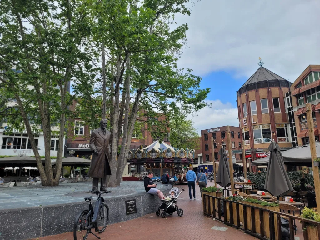 City Game Eindhoven lively square featuring the Frits Philips statue, surrounded by trees, charming cafes, and vibrant architecture, capturing the city's blend of culture and community spirit.